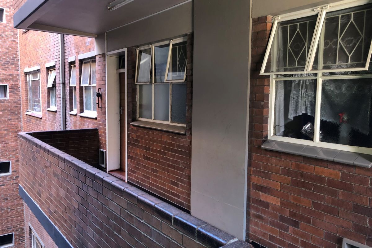 An exterior view of a brick apartment balcony featuring top-hung windows with security bars and a solid brick balustrade.