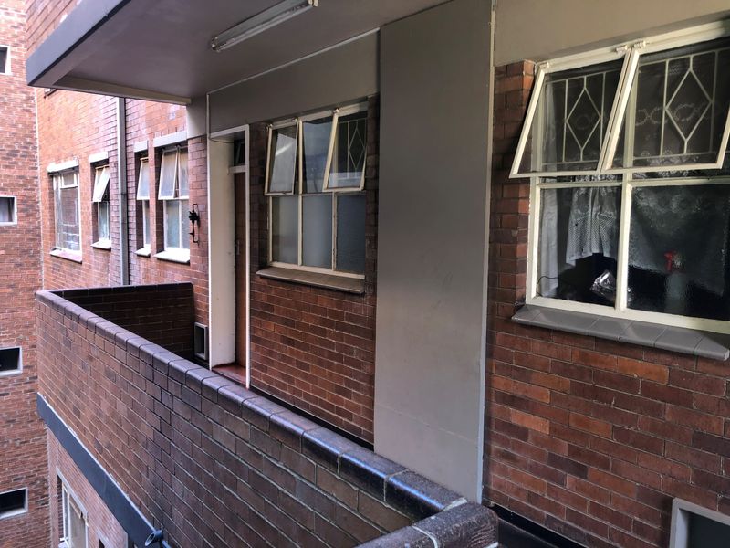 An exterior view of a brick apartment balcony featuring top-hung windows with security bars and a solid brick balustrade.