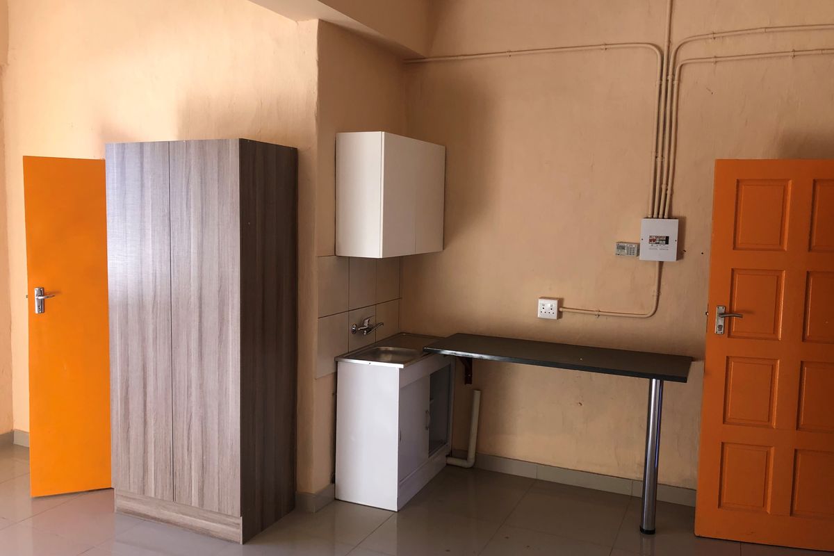 A compact kitchenette area featuring a small sink, base and wall cabinetry, and a simple countertop, set against a plain wall with exposed electrical conduits.