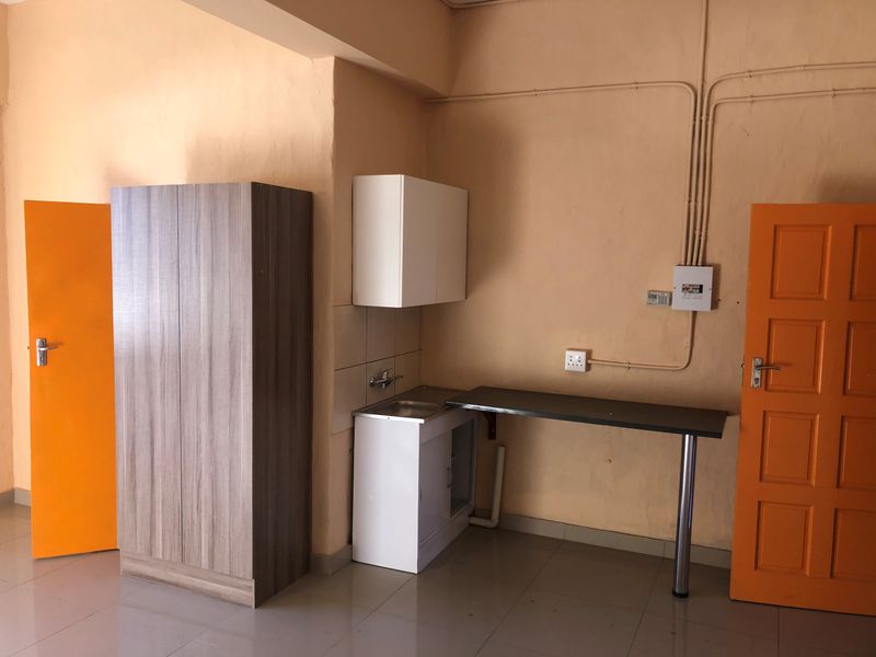 A compact kitchenette area featuring a small sink, base and wall cabinetry, and a simple countertop, set against a plain wall with exposed electrical conduits.