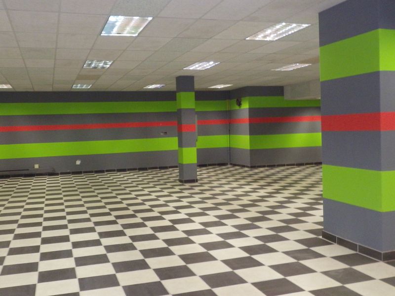 A large, open-plan commercial or utility space featuring a suspended ceiling, recessed lighting, and checkered floor tiles with structural columns.