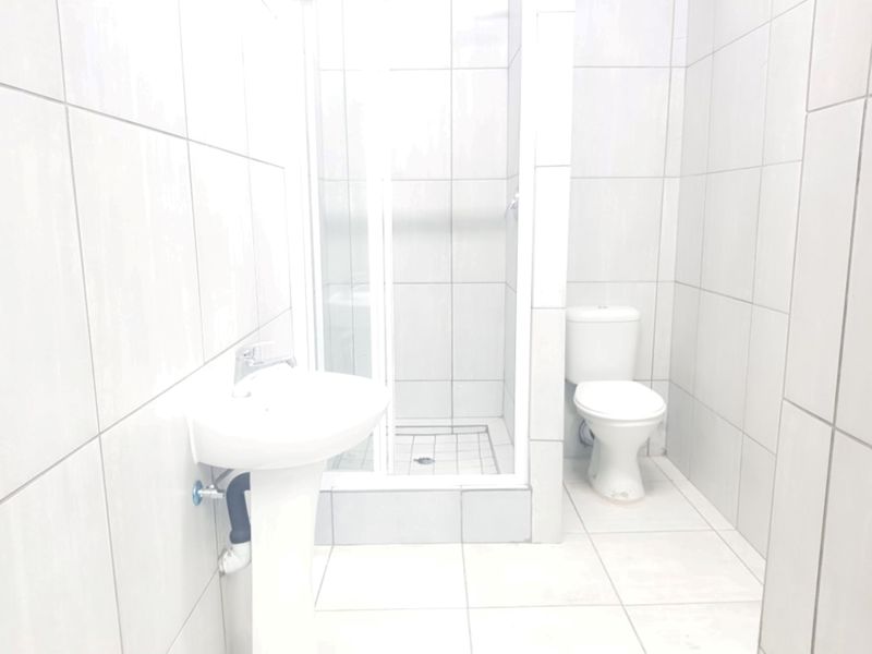 A functional bathroom featuring a pedestal washbasin, a shower enclosure, and a toilet, finished with neutral ceramic wall and floor tiles.
