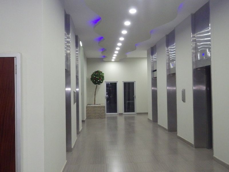 A modern, well-lit apartment building hallway featuring multiple elevators, tiled flooring, and a decorative recessed ceiling design.