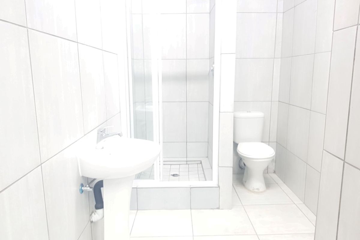 A functional bathroom featuring a pedestal washbasin, a shower enclosure, and a toilet, finished with neutral ceramic wall and floor tiles.