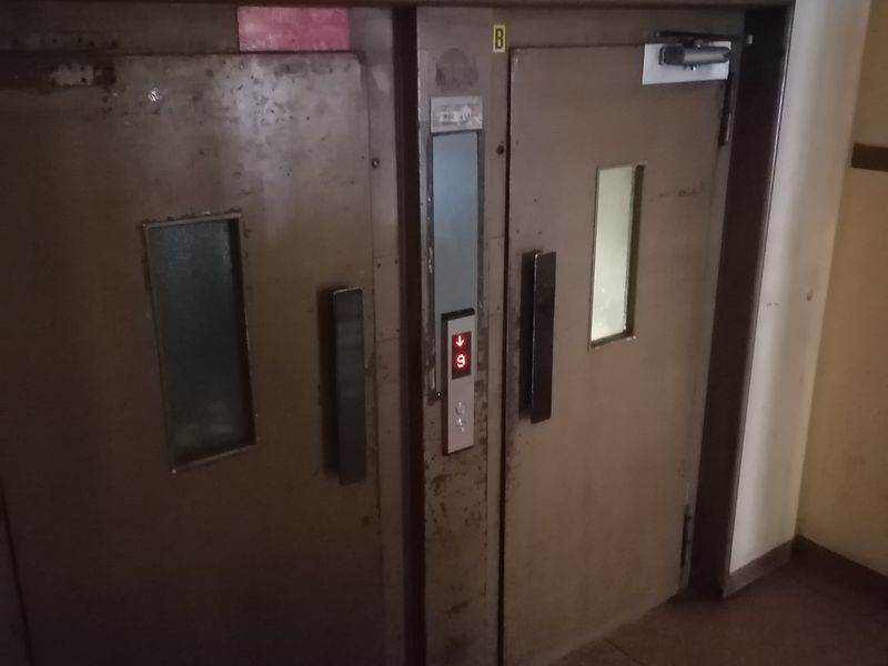 A view of the elevator lobby area featuring industrial-style metal elevator doors and a call panel in a dated hallway.