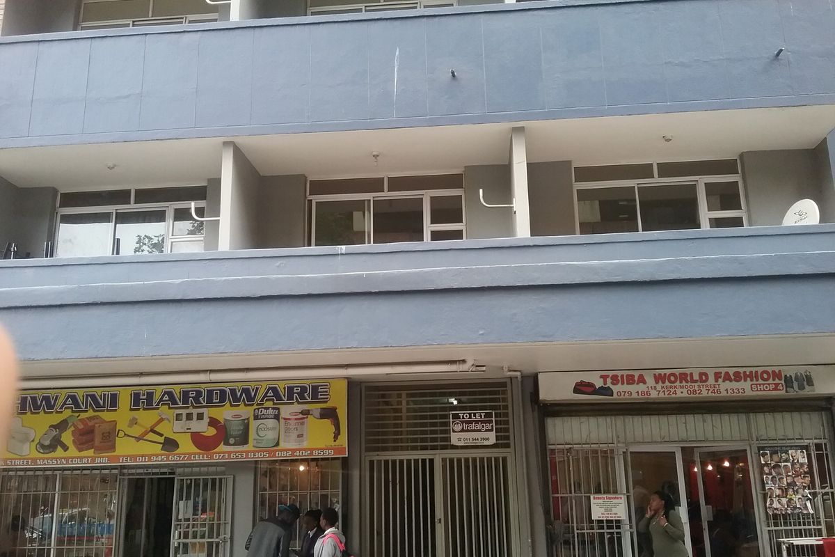 Street-level view of a multi-story commercial building featuring ground-floor retail storefronts with security gates and upper-level residential balconies.