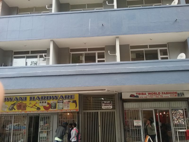 Street-level view of a multi-story commercial building featuring ground-floor retail storefronts with security gates and upper-level residential balconies.