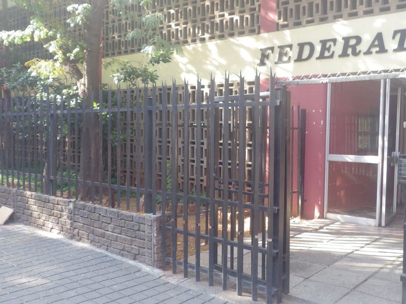 Front exterior view of the building entrance featuring a secure metal fence, brick boundary wall, and paved sidewalk access.