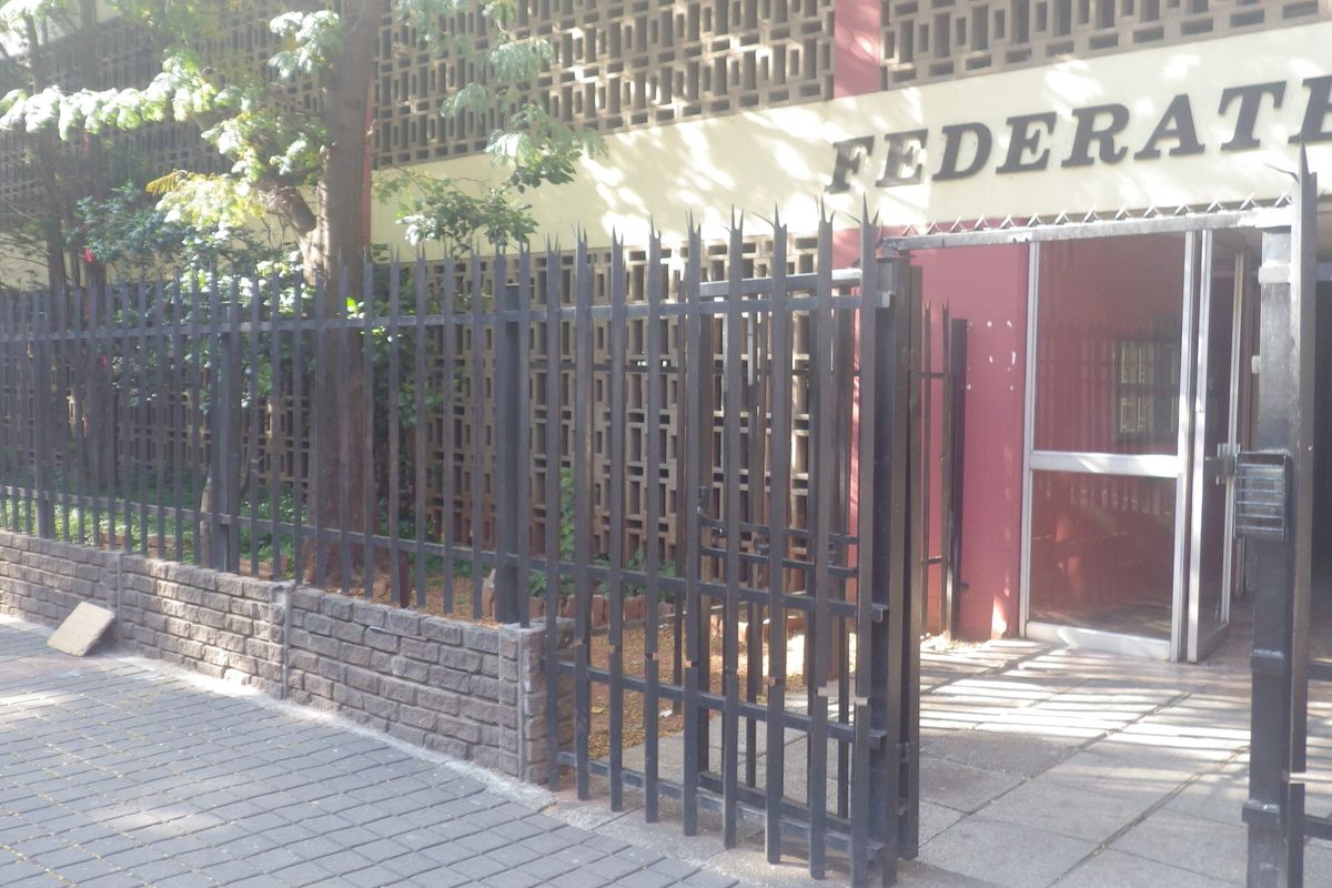 Front exterior view of the building entrance featuring a secure metal fence, brick boundary wall, and paved sidewalk access.