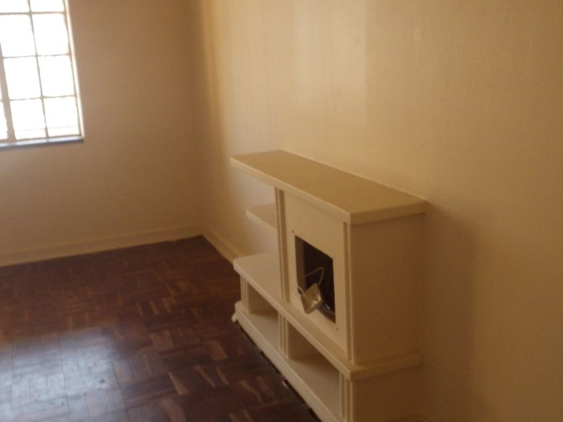 An empty living room featuring original parquet flooring, a grid-pattern window, and a built-in shelving unit requiring maintenance.
