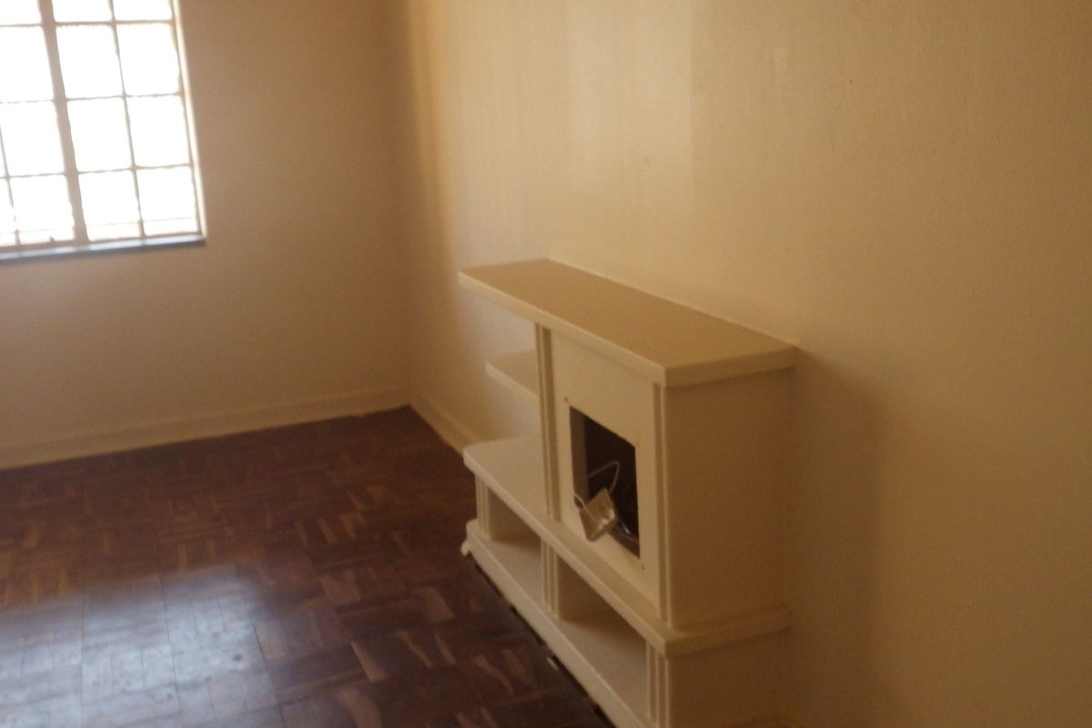 An empty living room featuring original parquet flooring, a grid-pattern window, and a built-in shelving unit requiring maintenance.