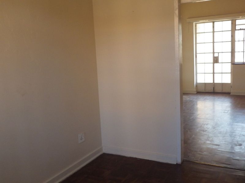 An empty living area featuring original parquet flooring and a doorway leading to a sunlit room with multi-pane glass doors.