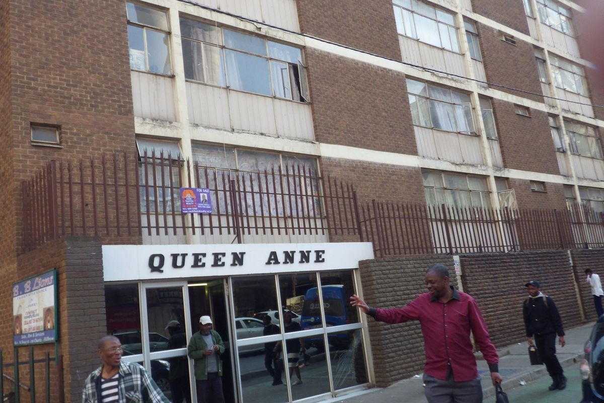 Street-level view of the Queen Anne apartment building facade, featuring brick construction, metal security fencing, and a glass-fronted entrance.