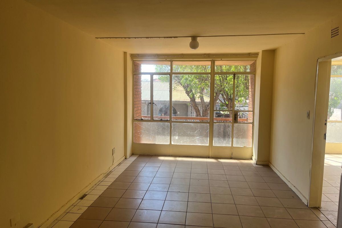 An empty, sunlit living area featuring tiled flooring and a large window with visible glass damage, offering a blank canvas for renovation.