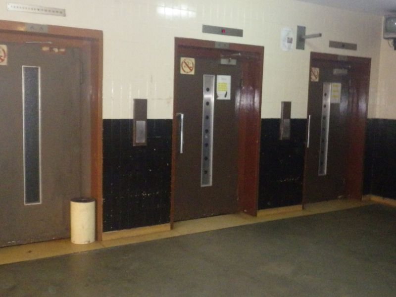 A communal hallway area featuring three elevator access points with dated wall tiling and flooring.