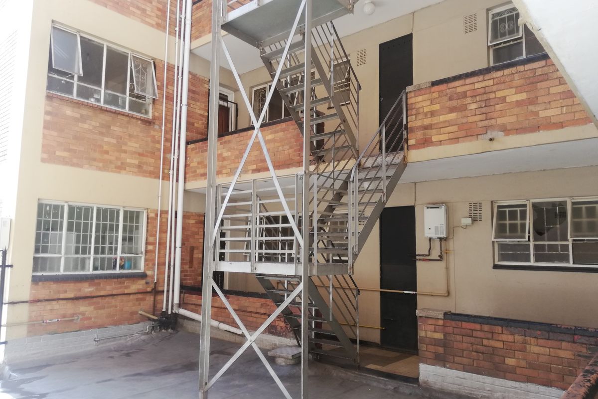 Rear exterior view of a multi-story flat building featuring a metal fire escape staircase, brick facade, and gas geyser installation.