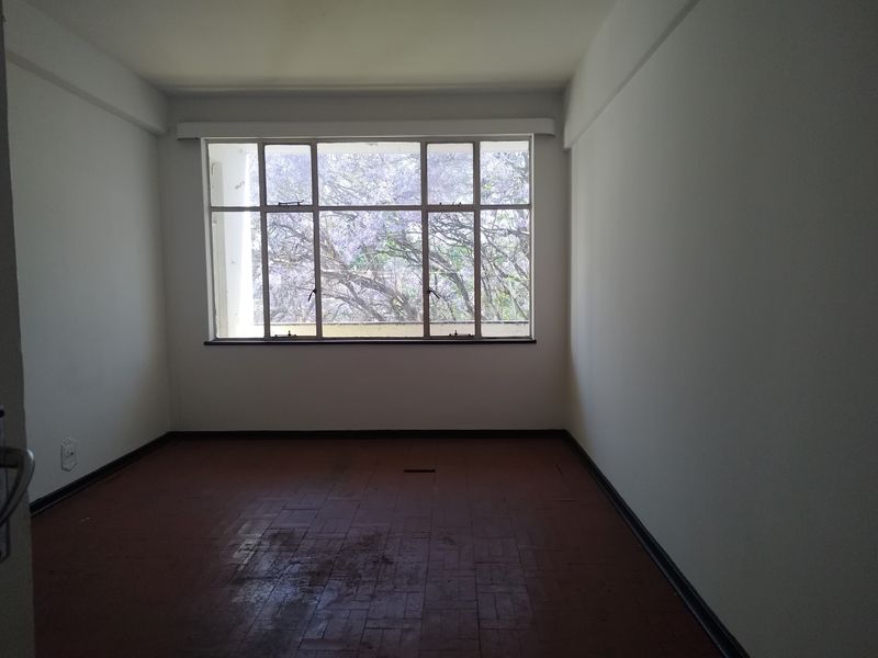 An empty bedroom featuring original parquet flooring and a large window providing natural light and a view of mature trees.