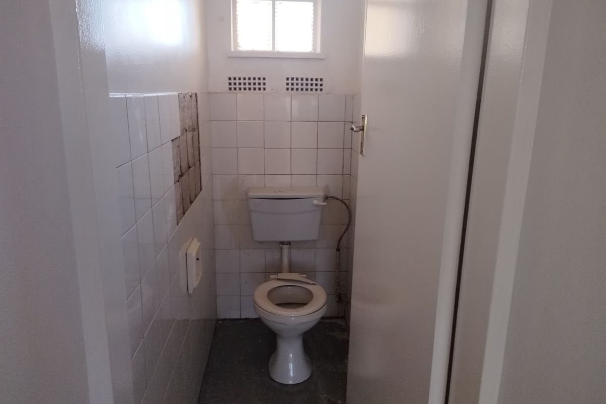 A small, dated bathroom featuring a standard toilet, partial wall tiling with visible missing tiles, and a small window for ventilation.