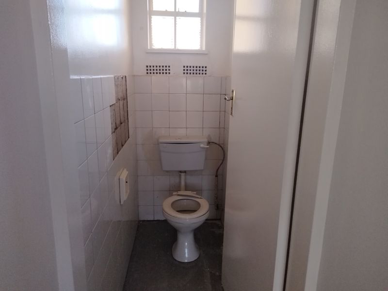 A small, dated bathroom featuring a standard toilet, partial wall tiling with visible missing tiles, and a small window for ventilation.