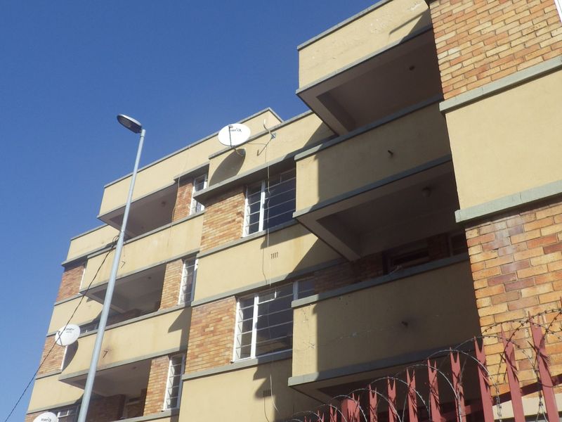 Exterior view of a multi-story brick apartment building featuring private balconies, steel-framed windows, and perimeter security fencing.