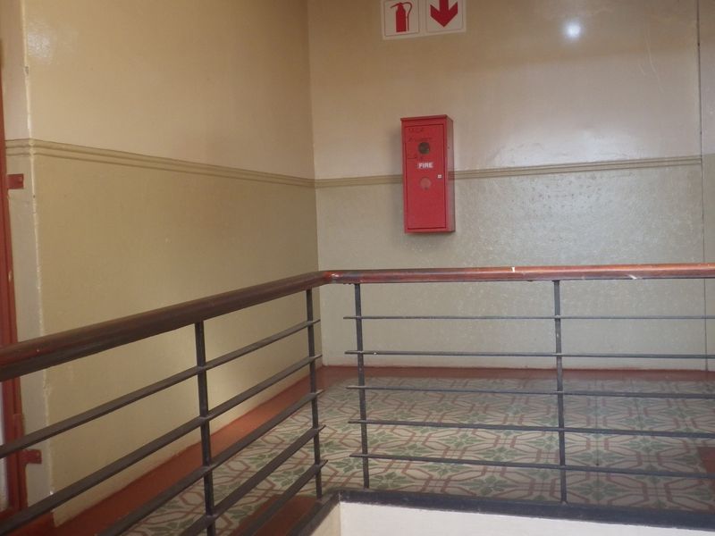 A communal hallway area featuring a metal balustrade with a wooden handrail, patterned floor tiles, and a wall-mounted fire hose reel.