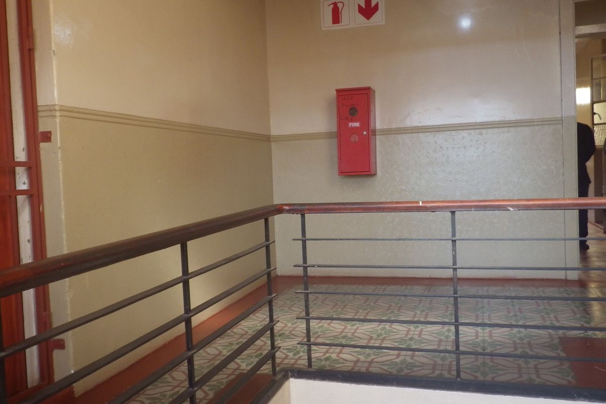 A communal hallway area featuring a metal balustrade with a wooden handrail, patterned floor tiles, and a wall-mounted fire hose reel.