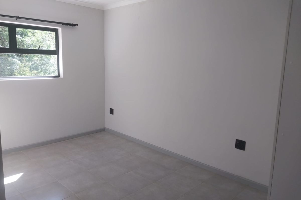 A clean, empty bedroom featuring neutral tiled flooring, modern grey skirting boards, and a window with a curtain rod.