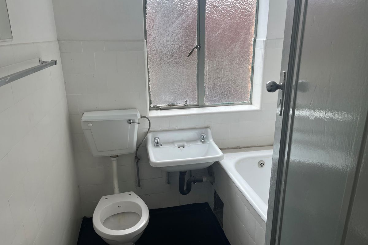 A compact, dated bathroom featuring a standard bathtub, wall-mounted basin, and toilet with white tiled walls and a window.