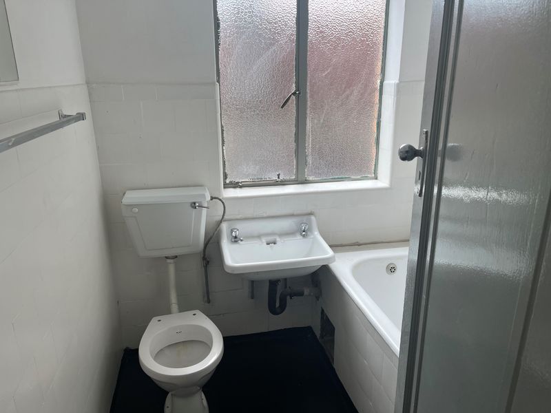 A compact, dated bathroom featuring a standard toilet, wall-mounted basin, and bathtub with tiled walls and a metal-framed window.