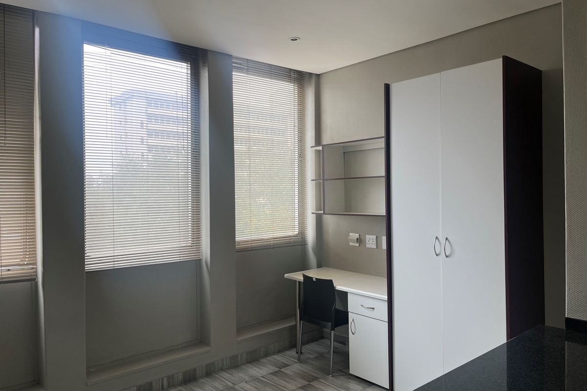 A modern bedroom featuring a built-in wardrobe, integrated desk with shelving, venetian blinds, and neutral tiled flooring.