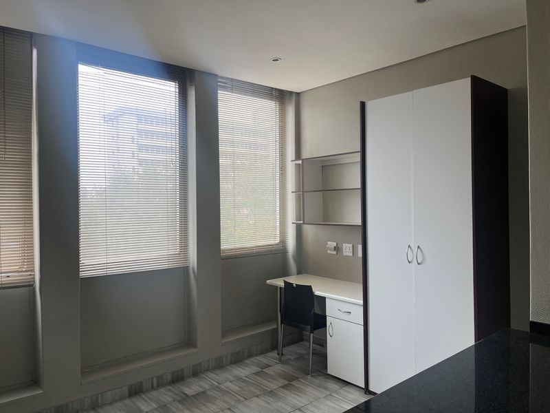 A modern bedroom featuring a built-in wardrobe, integrated desk with shelving, venetian blinds, and neutral tiled flooring.