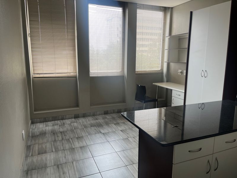 Open-plan living area featuring tiled flooring, built-in cabinetry with granite countertops, a dedicated desk space, and large windows with fitted venetian blinds.