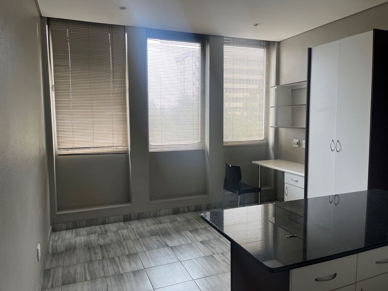 A modern bedroom featuring tiled flooring, built-in cupboards, a functional built-in desk, and large windows fitted with venetian blinds.