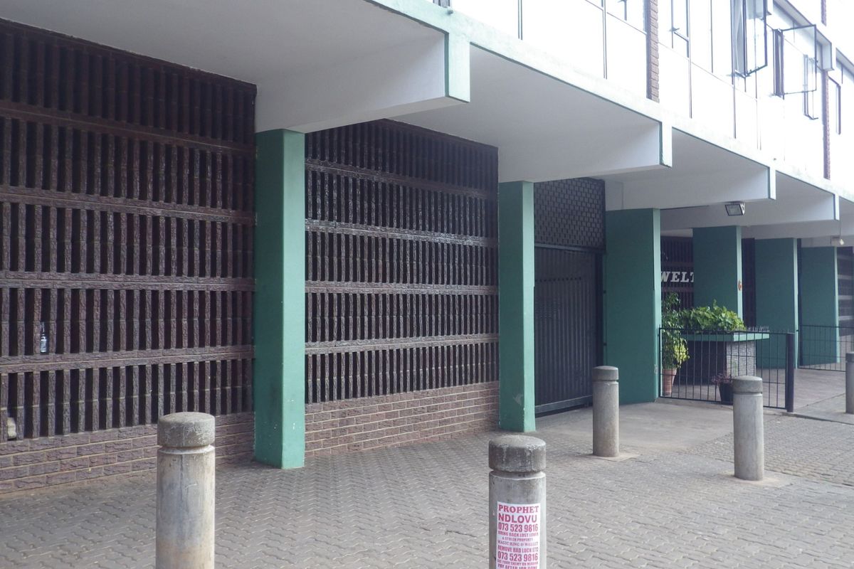 Exterior view of the building entrance featuring a brick screen wall, concrete pillars, and a paved walkway with security bollards.