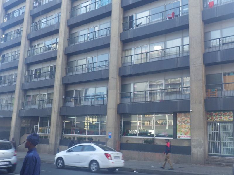 Street-level view of a multi-story apartment building featuring a concrete facade, private balconies with metal railings, and ground-floor commercial retail space.