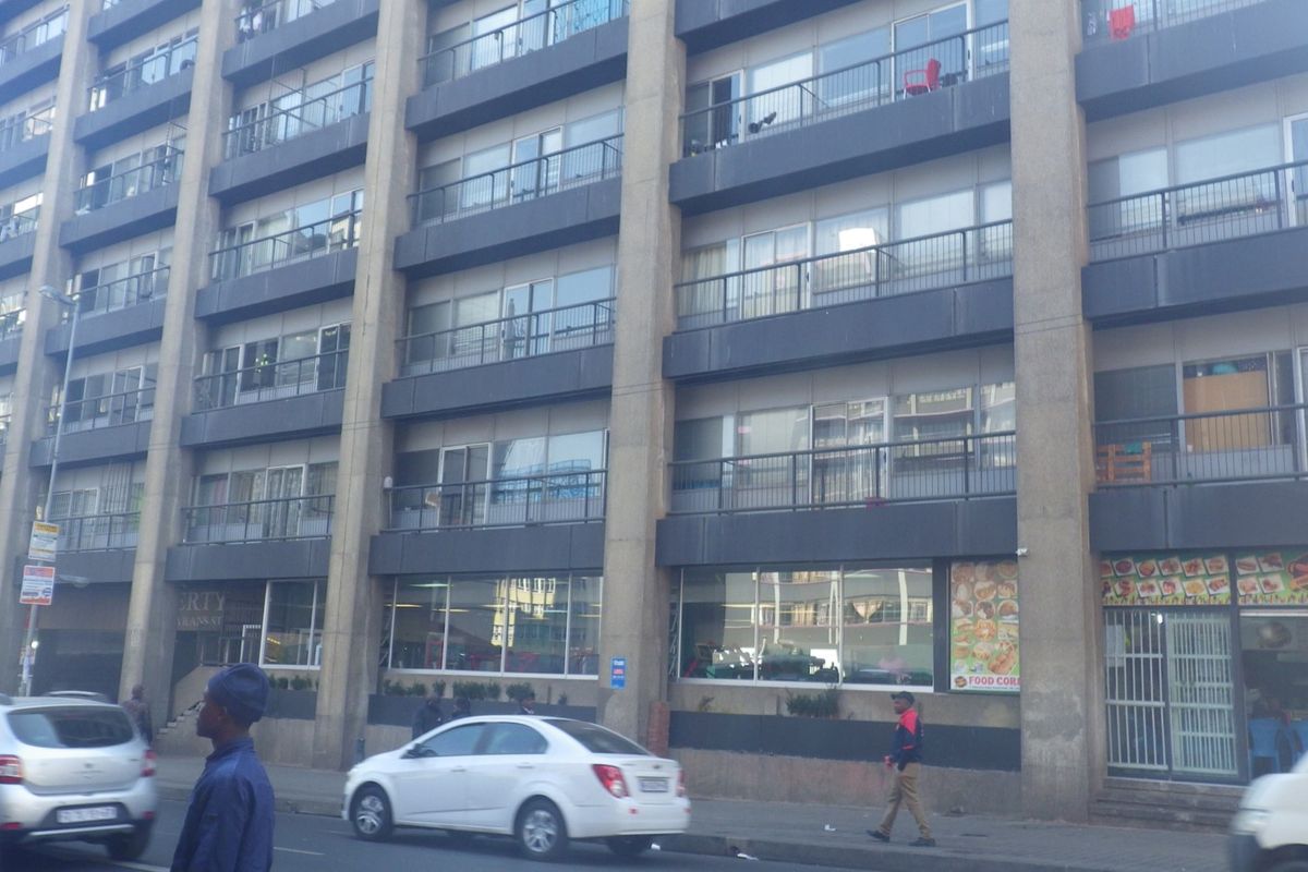 Street-level view of a multi-story apartment building featuring a concrete facade, private balconies with metal railings, and ground-floor commercial retail space.