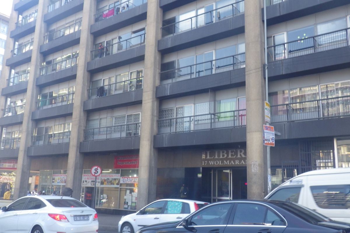 Street-level view of a multi-story apartment building featuring balconies, sliding doors, and ground-floor retail spaces in an urban setting.