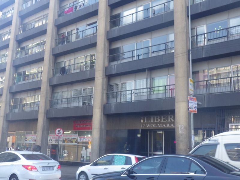 Street-level view of a multi-story apartment building featuring balconies, sliding doors, and ground-floor retail spaces in an urban setting.