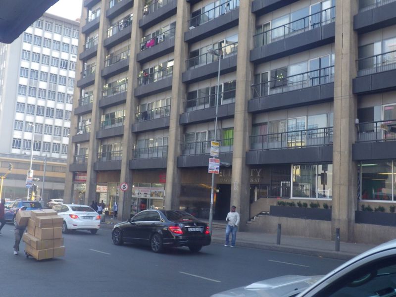 Street-level view of a multi-story apartment building featuring concrete architecture, private balconies, and ground-floor commercial space in an urban setting.