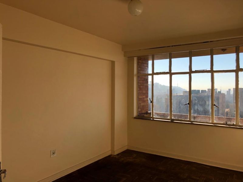 An empty bedroom featuring original parquet flooring, a large steel-framed window with city views, and exposed brick detailing.