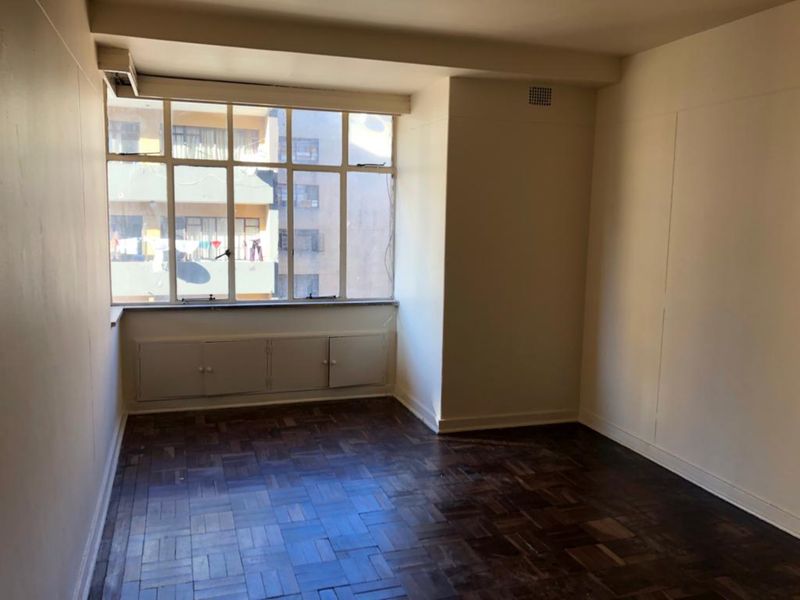 An empty living room featuring original parquet flooring, built-in under-window storage cabinetry, and a large window providing natural light.