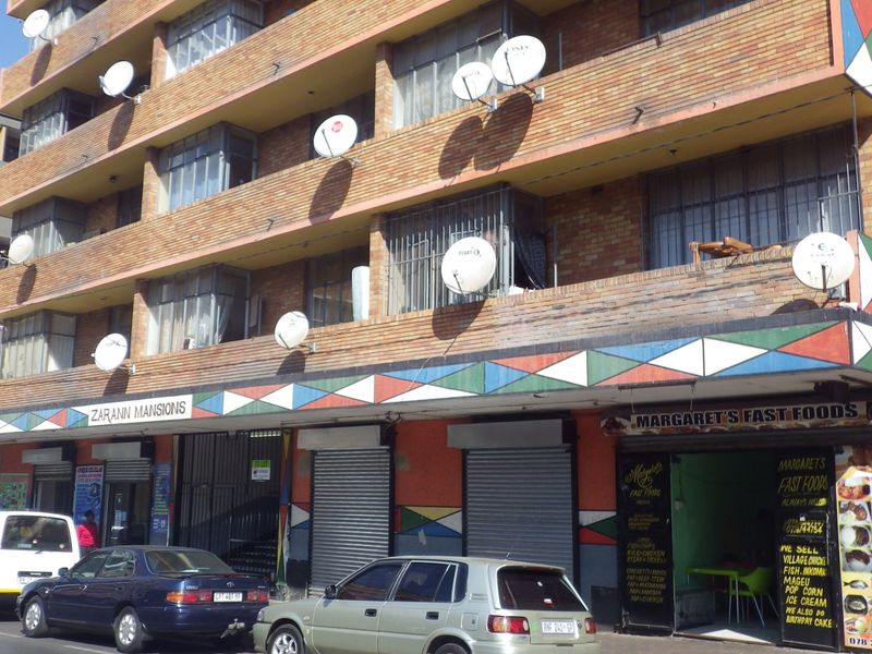 Street-level view of a multi-story brick apartment building featuring ground-floor commercial units, security shutters, and multiple satellite dishes on the facade.