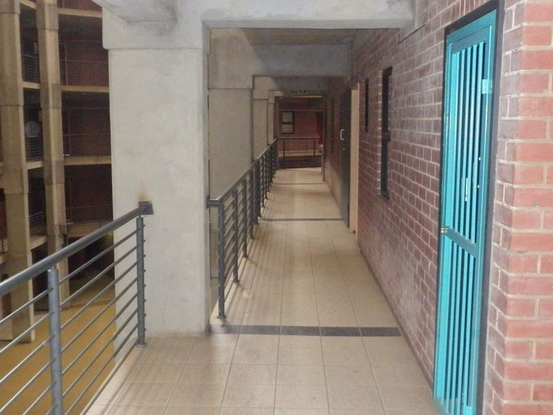 An open-air apartment complex hallway featuring exposed brick walls, tiled flooring, and metal safety balustrades overlooking the central courtyard.