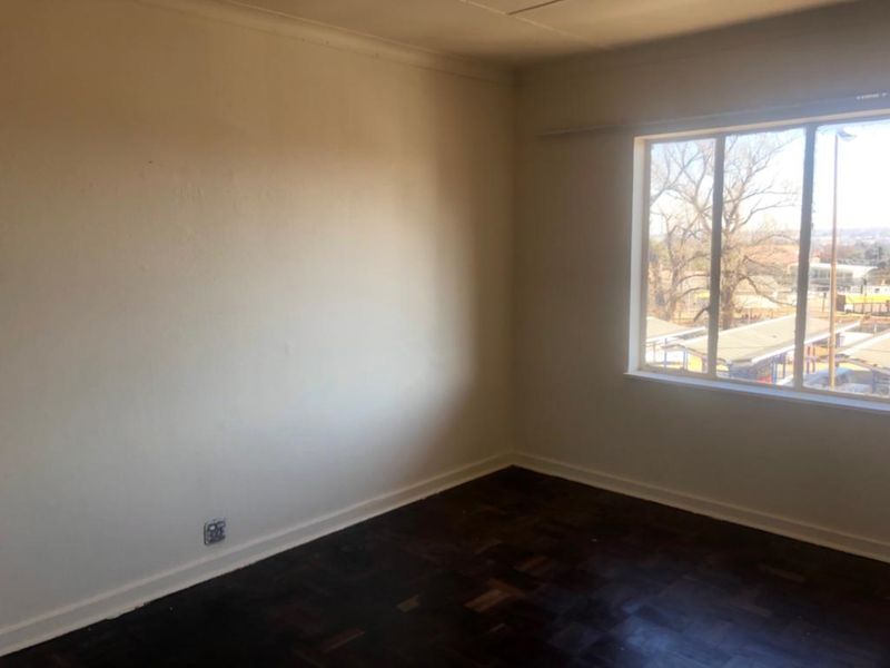 An empty bedroom featuring dark parquet flooring, neutral walls, and a large window providing natural light and an exterior view.