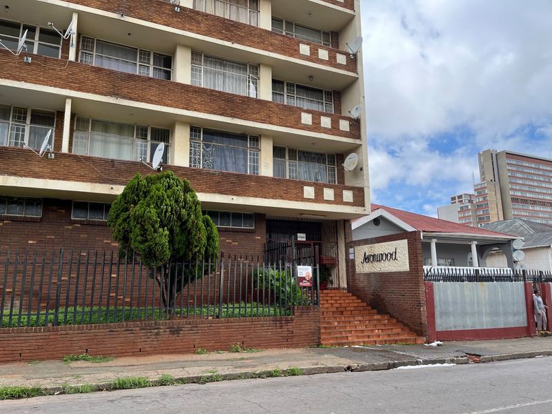 Street-level view of the Avonwood apartment building facade, featuring brick construction, security fencing, and a concrete staircase entrance.