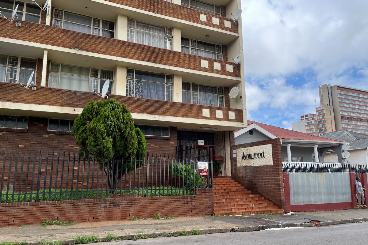 Street-level view of the Avonwood apartment building facade, featuring brick construction, security fencing, and a concrete staircase entrance.