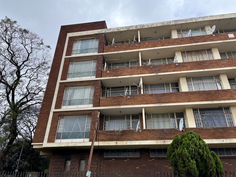 A multi-story brick apartment building featuring multiple balconies and steel-framed windows, showing signs of exterior weathering and maintenance requirements.