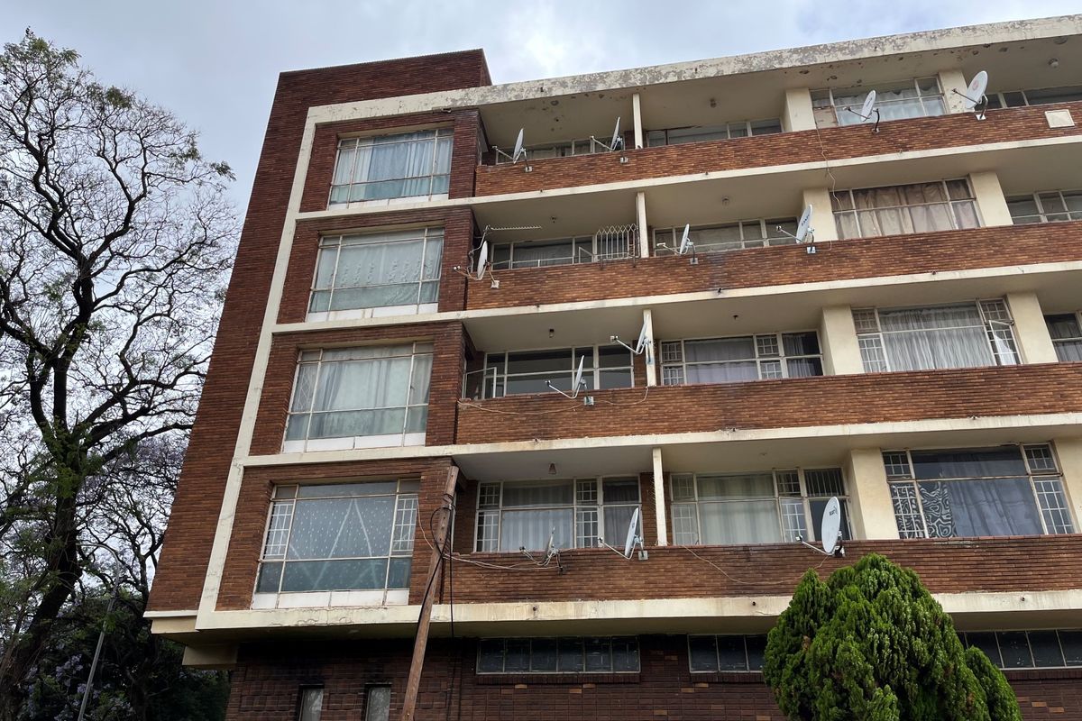 A multi-story brick apartment building featuring multiple balconies and steel-framed windows, showing signs of exterior weathering and maintenance requirements.