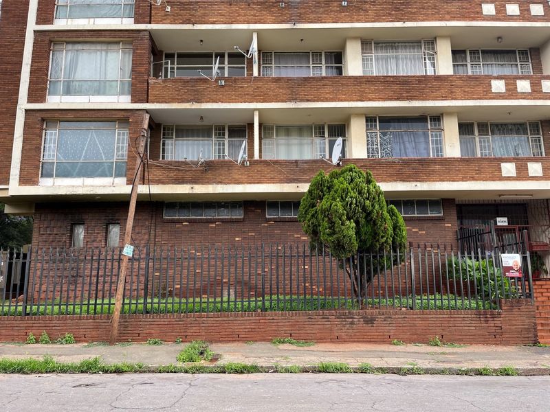 Front elevation of a multi-story brick apartment building featuring balconies, security fencing, and visible satellite dish installations.