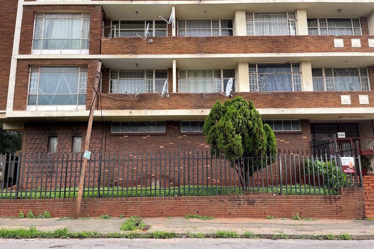 Front elevation of a multi-story brick apartment building featuring balconies, security fencing, and visible satellite dish installations.